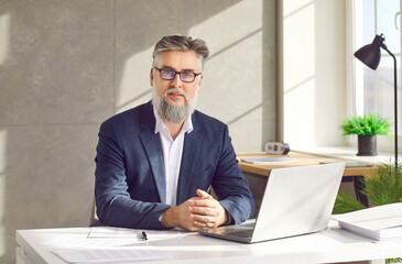 Portrait of a serious handsome bearded mature business man in suit and glasses working at the desk on his workplace with a laptop at office or at home and looking confident at the camera..