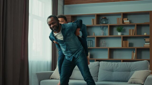 Happy family African American father dad man piggyback riding on back little funny son dancing with boy child kid play having fun at home cheerful dance playing together in cozy living room childcare