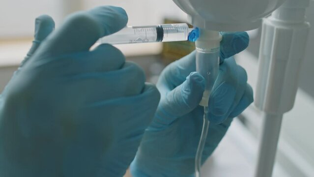 Close-up of unrecognizable nurse wearing latex gloves adding medication to IV bag for patient