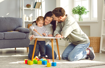Happy parents playing with their adorable baby at home. Mom and dad looking at their baby who drawing on paper with pencil in cozy living room. Creative education, early development
