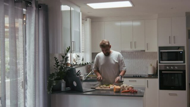 Young man watching tv show on computer and preparing dinner with vegetables in kitchen interior. Bearded man cooking while having a glass of wine