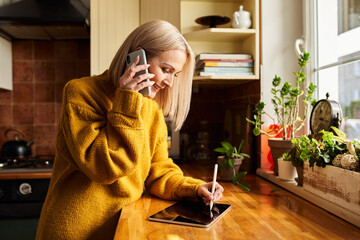 Adult woman writing on tablet while talking on the phone in a kitchen