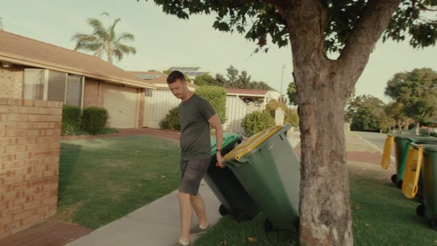 A man is hauling the household wheelie green and yellow bins for council gargbage collection recycable and organic waste from the street back home. Concept: Waste management