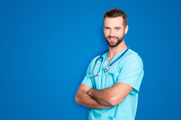 Portrait with copy space, empty place of positive cheerful man with stethoscope on his neck in blue lab uniform, having his arms crossed, isolated on grey background