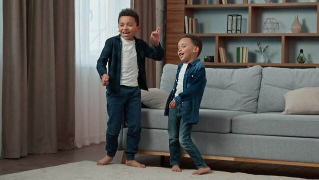 Two ethnic boys little children dancing to music in home living room. African American happy funny active moving multiethnic kids schoolboys pupils brothers dance to song watching TV move having fun