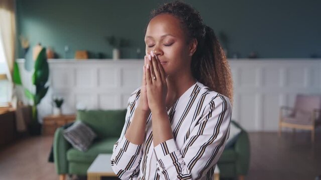 Young unhappy ethnic African American woman nervous and stressed when hears about occurrence of difficult situation and tries to pray in hope of good luck, stands in spacious living room