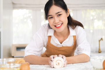 Obraz na płótnie Canvas Asian beautiful woman stay home, spend time in kitchen cooking. Young attractive carrying female wear apron and smile, holding yeast dough with hand to baking bakery on table for dinner in house.