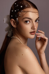 Female model with red hair, brown eyes, freckled skin, poses for a beauty shoot. Her hair is in a ponytail. She has a white bow in her hair. She has pearls in her hair. Model has her hand to her face.