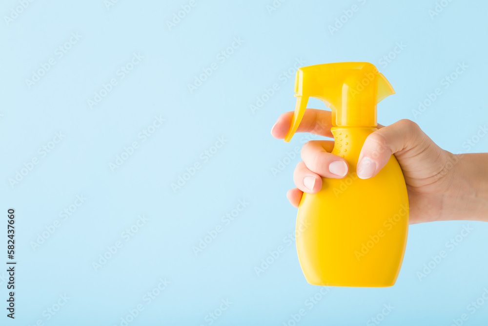 Wall mural Young adult woman hand holding yellow spray bottle of sunscreen or cleaning detergent on light blue wall background. Pastel color. Closeup. Empty place for text. Side view.