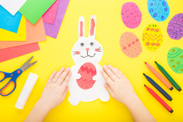 Little child hands with white smiling bunny and colorful eggs from paper on bright yellow table...