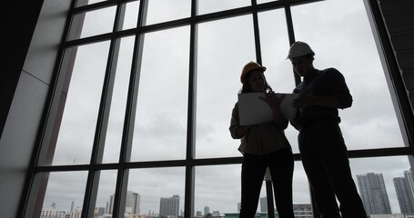 Fototapeta na wymiar Two architect and engineer working on plans with blueprint standing near length window at cityscape with skyscrapers