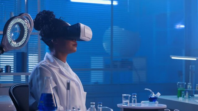 African American woman in virtual reality headset examines the sample, touches, zooms, scrolls the image. Black female doctor or researcher works in a biochemical modern laboratory with blue light.