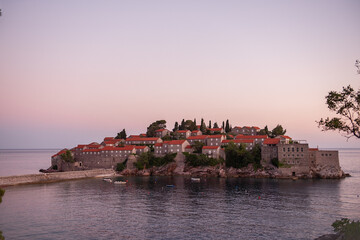 Naklejka premium Panoramic view of Sveti Stefan in Montenegro at pink sunset. Famous tourist place near Budva. Natural beautiful island, with terracotta roofs among the turquoise sea. Copy space, wallpaper background