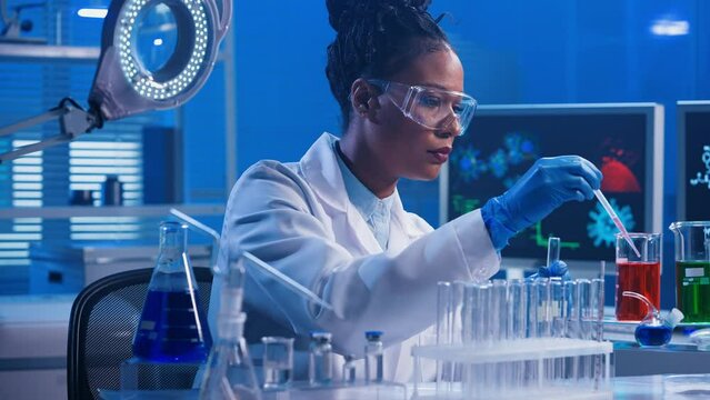 A young African American woman using a plastic pipette pipettes red and green liquid into glass test tubes and examines samples. A black female doctor works in a biochemical laboratory with blue light