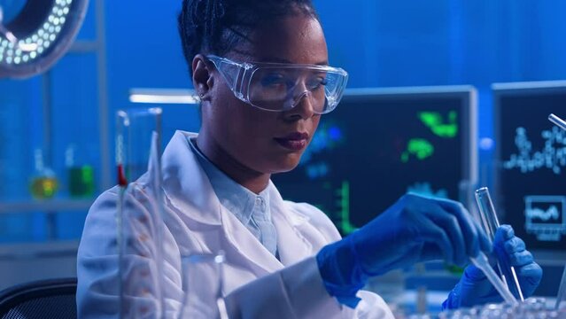A young African American woman using a plastic pipette pipettes red liquid into glass test tube and examines sample. A black female doctor works in a biochemical laboratory with blue light. Close up.