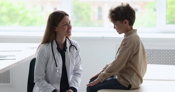 Engaged caring pretty female pediatrician with stethoscope talking to little patient boy, asking about complaints, speaking, smiling, laughing, examining child in doctor office