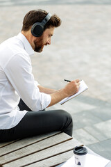 Serious manager in headphones writing in diary while sitting
