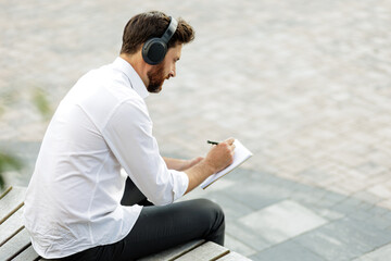 Focused manager in headphones writing in notepad outdoors