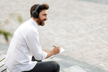 Fototapeta na wymiar Man in headphones sitting outdoors and writing in diary