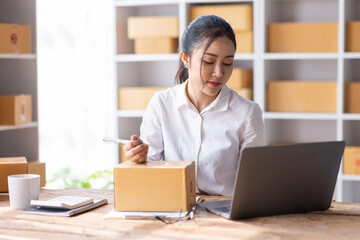 Fototapeta premium Portrait of Starting Asian female small businesses owners SME entrepreneur working at Laptop in home Warehouse, online marketing, packing boxes, SME sellers, concept, e-commerce team, online sales.
