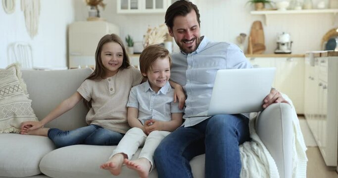 Happy daddy and two cute little sibling kids using online app on laptop for communication, watching cartoon funny movie, sitting with laptop on comfortable sofa