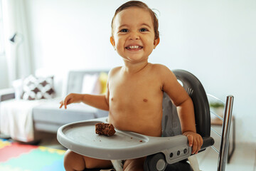 Portrait of a beautiful and adorable child sitting in a high chair eating homemade cake. Everyday...