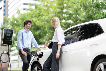 Progressive businessman and businesswoman with electric car parking and connected to public...
