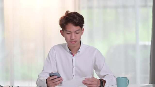 Front view of young asian man in white shirt using mobile smart phone for paying electricity bills at desk