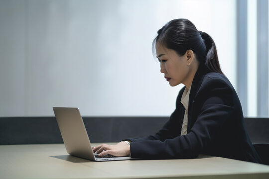 A Asian business woman sits at a desk in a suit and is typing on a laptop. Generative AI