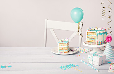Birthday party table setting with slice of vanilla confetti cake and decorations on a light grey...