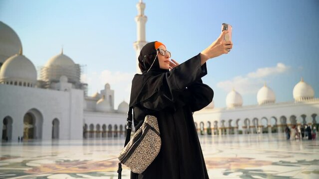 Woman in hijab takes selfie in front of mosque in UAE spbas. Young beautiful female holding smartphone in hand and taking photo, looking with smile and standing against background of white