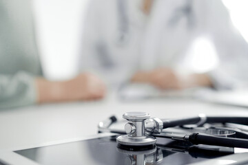 Doctor and patient are sitting and discussing something at the desk in the clinic office. The focus is on the stethoscope lying on the table, close up. Perfect medical service and medicine concept