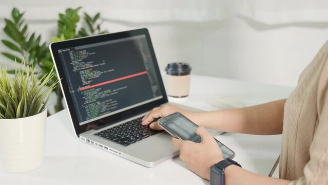 Programmer woman looking mobile phone program code at workplace, software developer hands typing data coding programming javascript with laptop computer, IT startup concept, Web application design