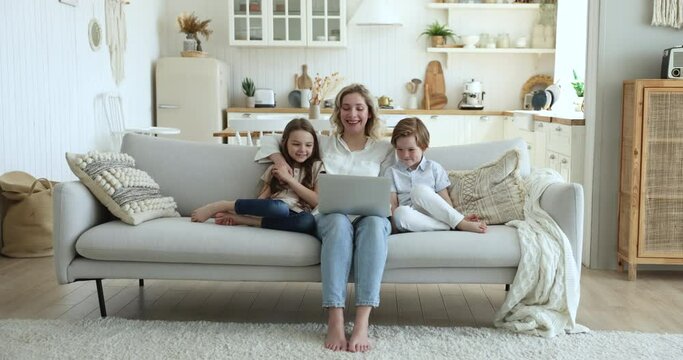 Cheerful beautiful millennial mom and two sweet little kids watching movie, cartoon on laptop together, resting on sofa at home, looking at display, smiling, laughing, Full length