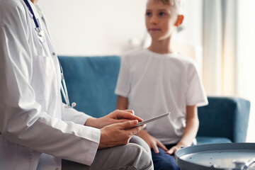 Obraz premium Doctor woman and kid boy patient at home. The pediatrician using tablet computer while filling up medical records, close up. Medicine, healthcare concepts