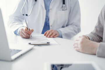 Doctor and patient sitting at the table in clinic. The focus is on female physician's hands filling up the medication history record form or checklist, close up. Medicine concept