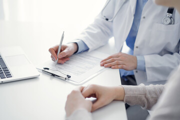 Doctor and patient sitting at the table in clinic. The focus is on female physician's hands filling up the medication history record form or checklist, close up. Medicine concept