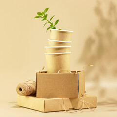 Sustainable still life. Eco friendly packaging concept. Cardboard boxes, disposable cups with green shoot on beige background. Concept of ecology, conservation, plastic free and recycling. Front view