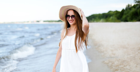 Fototapeta na wymiar Happy smiling woman in free happiness bliss on ocean beach standing and posing with hat and sunglasses. Portrait of a female model in white summer dress enjoying nature.
