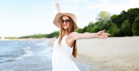 Fototapeta na wymiar Happy smiling woman in free happiness bliss on ocean beach standing with a hat, sunglasses, and rasing hands. Portrait of a multicultural female model in white summer dress enjoying nature.