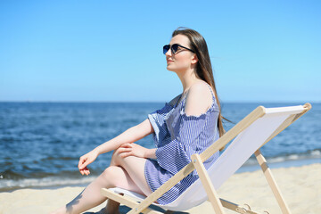 Happy young brunette woman relaxing on a wooden deck chair at the ocean beach while smiling, and wearing fashion sunglasses. The enjoying vacation concept