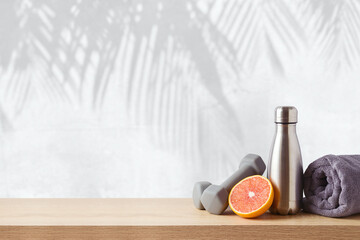 Summer fitness background with bottle of water, orange and dumbbells on wooden table over wall with palm tree shadows - 582527499