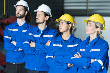 Group of male and female engineer workers wearing safety uniform and helmet standing together, looking to the workshop and crossed arms at production line in industry factory