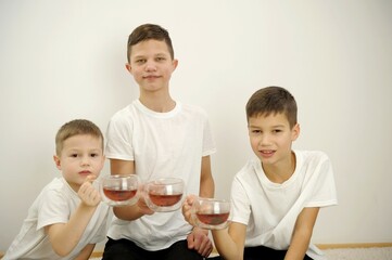 three boys on white background drink tea from glass mugs. They look into frame different ages 5 years old 9 years old 13 years old White t-shirts brothers similar eyes white background teenager