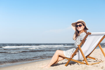Obraz premium Happy brunette woman wearing sunglasses and hat relaxing on a wooden deck chair at the ocean beach