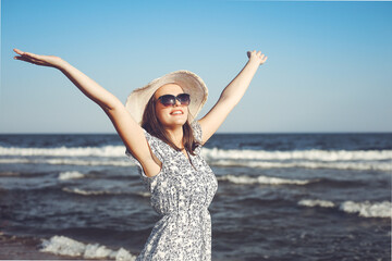 Fototapeta na wymiar Happy brunette beautiful woman on the ocean beach standing in a white summer dress and glasses, raising hands