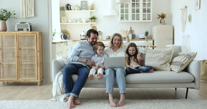 Positive mom, dad, two little kids watching funny cartoon movie on couch, making family video call, enjoying domestic internet online communication, smiling, laughing, Full length