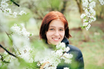 A teenage girl smiles in front of blooming tree in spring, she gathers positive energy and strength after a long winter, the girl is happy, her spring depression has already gone