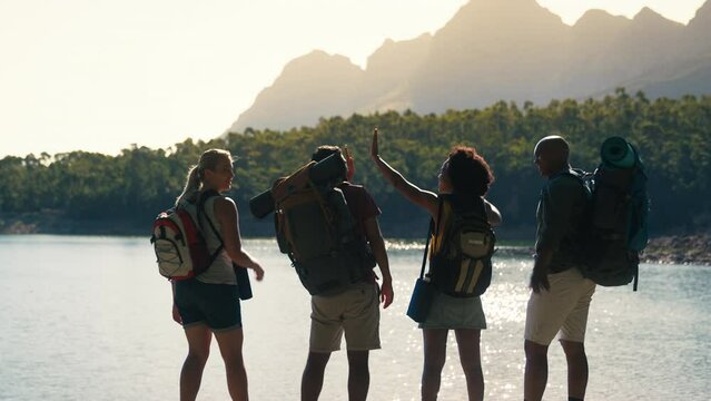 Rear view of group of friends with backpacks on hiking vacation in countryside looking out over beautiful lake and mountains giving each other high five - shot in slow motion