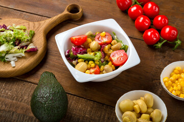 Vegetable salad with champignon corn and cherry tomato with avocado and olive oil in a white plate on a wooden table next to all the ingredients.